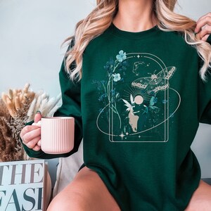 May include: Dark green sweatshirt featuring a nature-themed graphic. The design includes a butterfly, flowers, a fairy, and celestial elements within a rectangular frame. The sweatshirt is a solid dark green color.