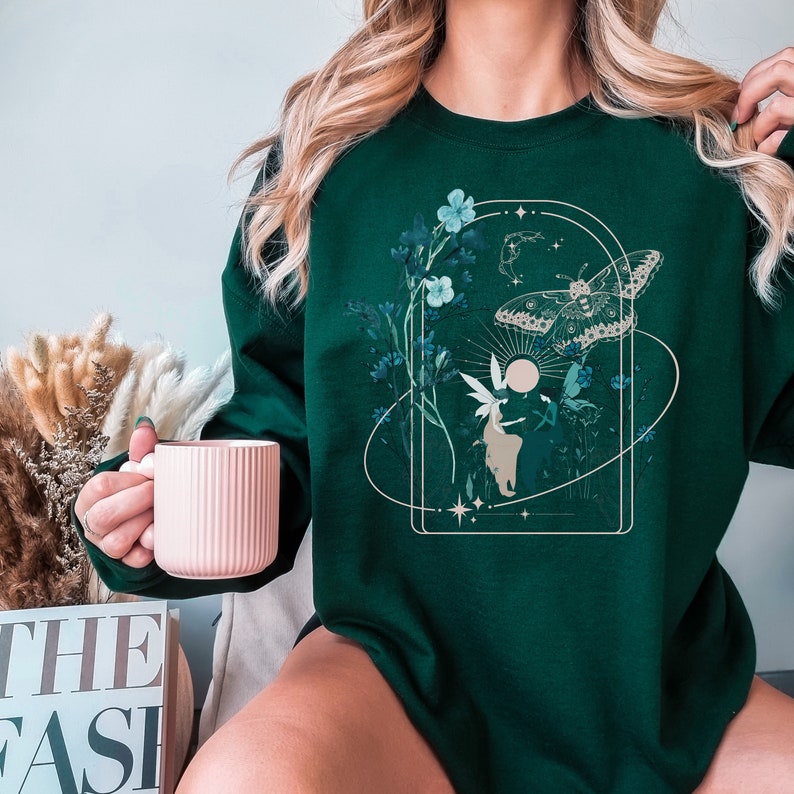 May include: Dark green sweatshirt featuring a nature-themed graphic. The design includes a butterfly, flowers, a fairy, and celestial elements within a rectangular frame. The sweatshirt is a solid dark green color.