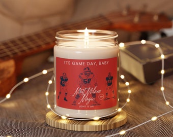 May include: A lit glass jar candle with a red label that reads "IT'S GAME DAY, BABY." The label features football player illustrations and the text "Matt Milano Magic." The candle sits on a wooden coaster, with string lights and a guitar in the background.