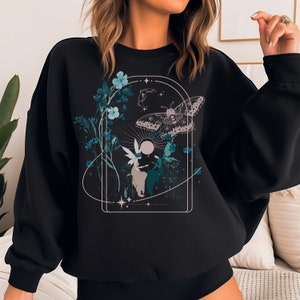 May include: Black sweatshirt featuring a nature-themed graphic. The design includes a moth, fairies, flowers, and celestial elements in teal, white, and beige. The artwork is framed within an arched border, creating a whimsical and ethereal aesthetic.