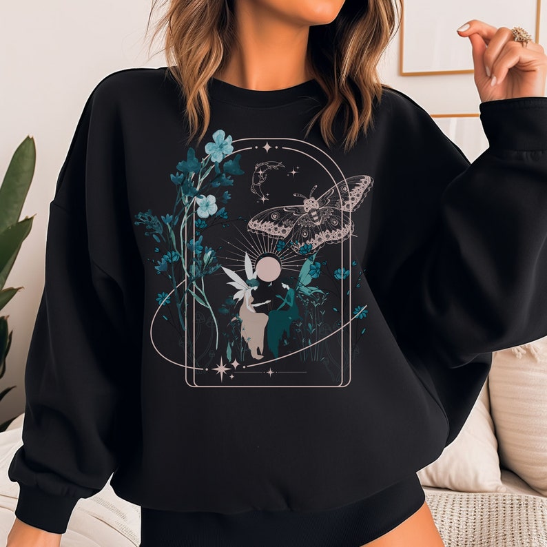May include: Black sweatshirt featuring a nature-themed graphic. The design includes a moth, fairies, flowers, and celestial elements in teal, white, and beige. The artwork is framed within an arched border, creating a whimsical and ethereal aesthetic.