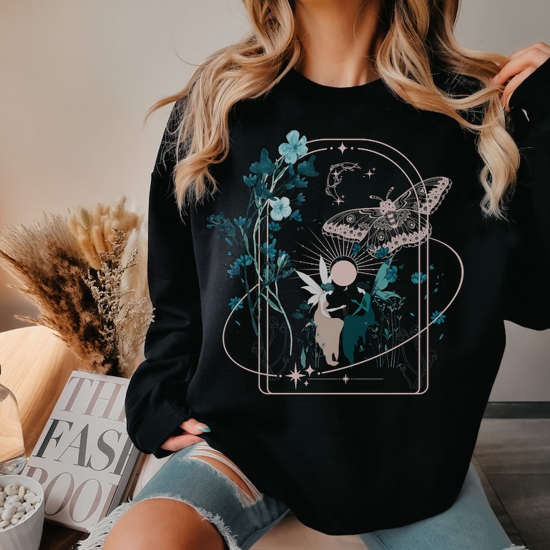 May include: Black sweatshirt featuring a nature-themed graphic. The design includes a butterfly, flowers, and a fairy-like figure within a geometric frame. The artwork is in shades of teal, beige, and white.