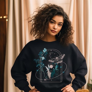 May include: Black sweatshirt featuring a nature-inspired graphic. The design includes a butterfly, flowers, and celestial elements within an arched frame. The artwork is in shades of teal, white, and beige.