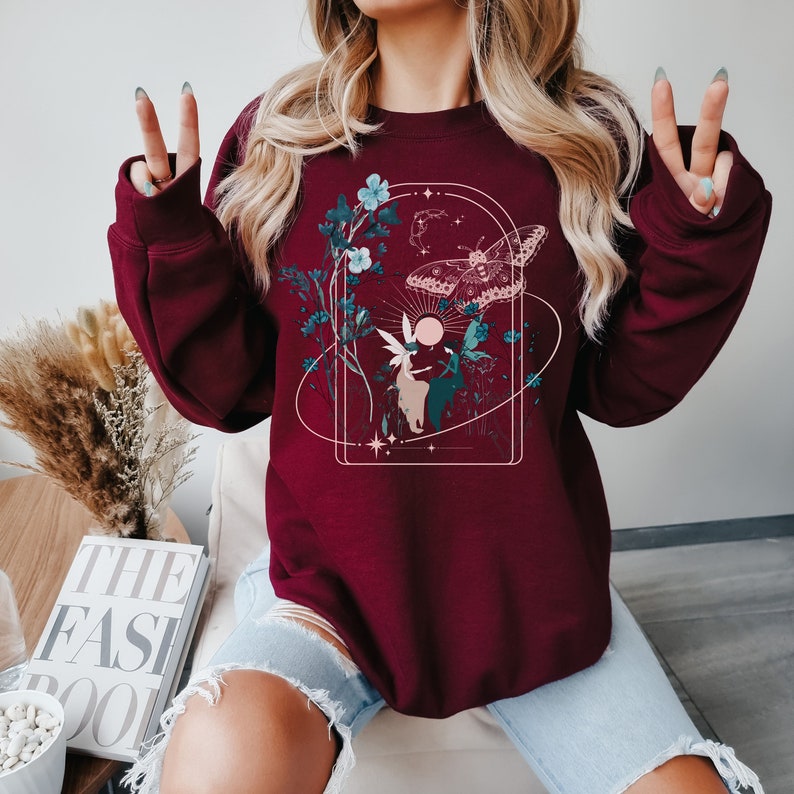 May include: Burgundy sweatshirt featuring a nature-themed graphic. The design includes a butterfly, flowers, and a fairy within an arched frame. The artwork is in shades of teal, beige, and white. The sweatshirt is a solid burgundy color.