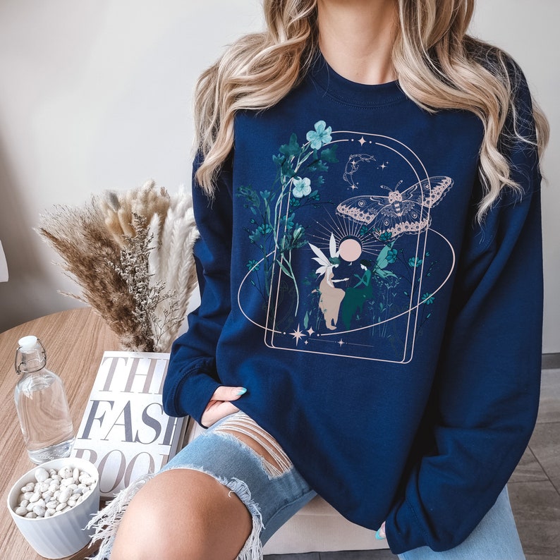 May include: Navy blue sweatshirt featuring a nature-themed graphic. The design includes a butterfly, flowers, and a fairy within a gold-colored arched frame. The sweatshirt is a long-sleeved crew neck.