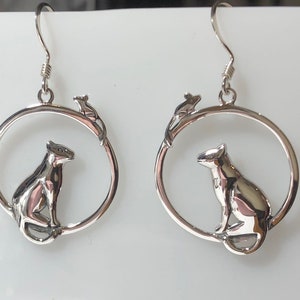 May include: Silver earrings with a cat and a mouse design. The cat is sitting inside a circle and the mouse is on a branch above the cat.