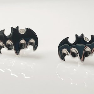 May include: Two silver-toned Batman logo stud earrings. Each earring features a black enamel bat silhouette with a glossy finish. The earrings are displayed on a white surface, reflecting the light.