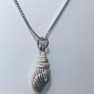 May include: A silver seashell pendant necklace. The pendant features a detailed, spiralled seashell design with a small, light blue gemstone. The necklace chain is a delicate box chain style. The jewellery is displayed against a white background.