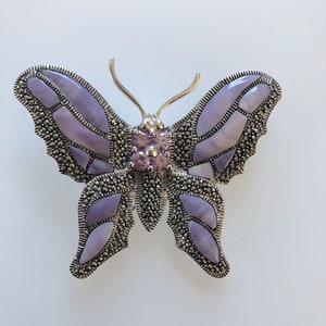 Puede incluir: Un broche de mariposa de plata con alas de concha de abulón púrpura y detalles de cristal rosa.