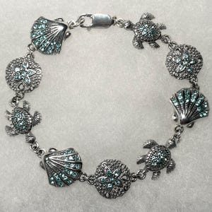 May include: Silver-toned bracelet featuring sea-themed charms. The bracelet includes seashell, starfish, and turtle charms, each adorned with small, light blue crystals. The bracelet is connected by a chain and has a clasp closure.