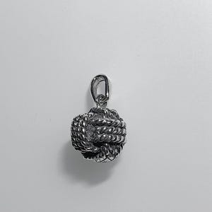 May include: A silver pendant featuring a detailed knot design. The pendant has a small loop for a chain and a textured surface. The knot is round and intricate, with a small, sparkling accent in the centre. The pendant is silver in colour.