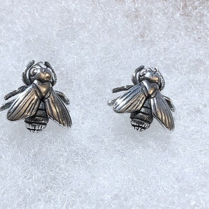 May include: A pair of silver-colored bee-shaped stud earrings. The earrings feature detailed wings, antennae, and body segments. The earrings are displayed on a white, textured surface.