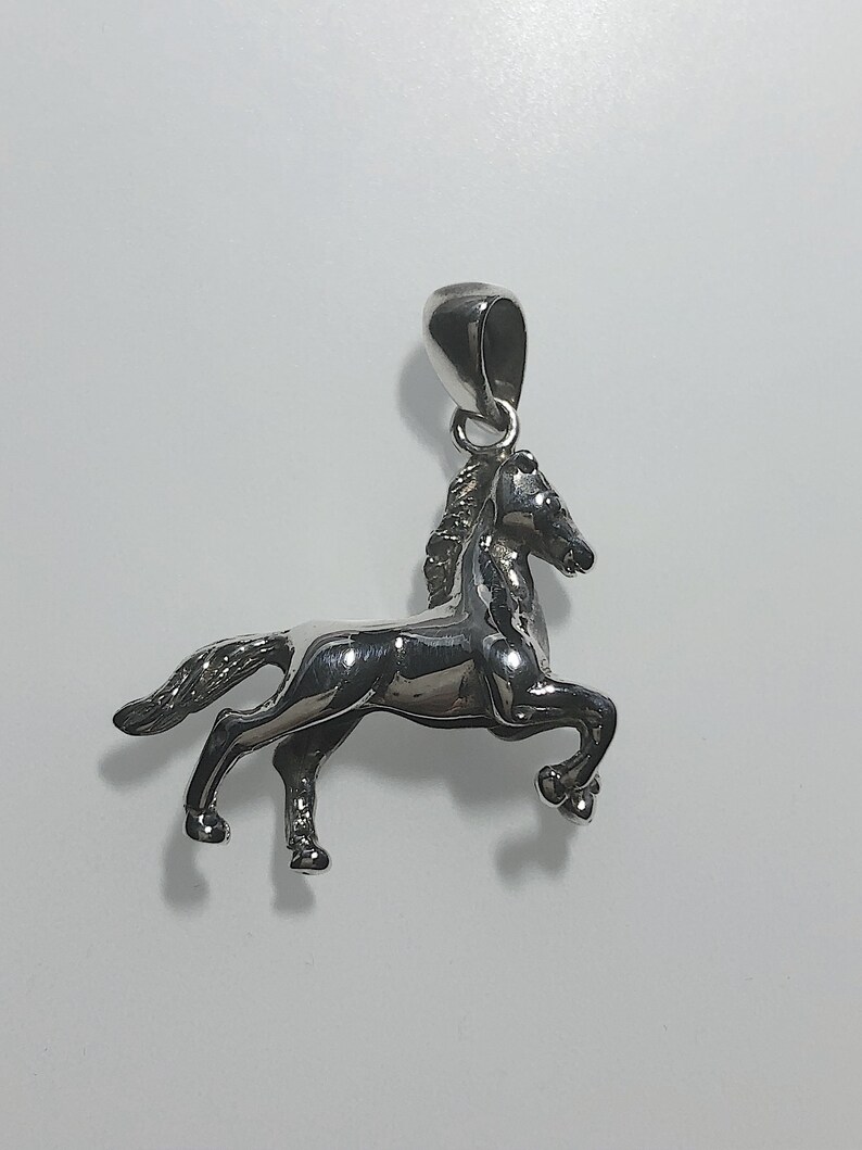 May include: Silver horse pendant charm. The horse is standing on its hind legs with its head turned to the side. The pendant has a loop at the top for attaching to a chain.