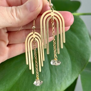 May include: Gold-colored earrings with a geometric arch design. Each earring features a series of vertical bars and a small chain that suspends a clear, faceted crystal. The earrings are displayed against a green leafy background.