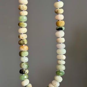 May include: A necklace featuring a strand of oval-shaped beads in shades of green, cream, and yellow. The beads are strung together with a thin, light-colored cord. The necklace is displayed against a gray background.