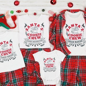 May include: A set of four white t-shirts with red and black text that reads "Santa's Cookie Crew" with different roles for each shirt: Taste Tester, Chief Decorator, Head Baker, and Bakers Assistant. The shirts are paired with red and green plaid pajama pants.