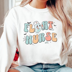 May include: White long-sleeved sweatshirt with a graphic design of a ghost wearing a nurse's hat and holding a syringe. The text "FLOAT NURSE" is written in a retro font with colorful letters.