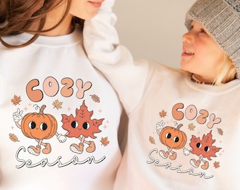 May include: White sweatshirts with a pumpkin and a maple leaf design. The text "Cozy Season" is printed on the sweatshirts.