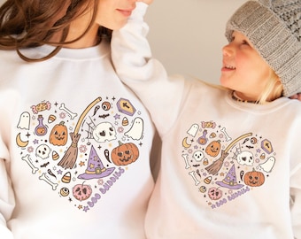 May include: Two white sweatshirts with a heart-shaped design featuring various Halloween-themed illustrations, including pumpkins, ghosts, skulls, a witch's hat, and a broom. The text "Boo Buddies" is printed on both shirts.