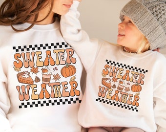May include: Two white sweatshirts with the text "Sweater Weather" in a retro font. The sweatshirts are decorated with fall-themed illustrations, including pumpkins, leaves, coffee cups, and pie.