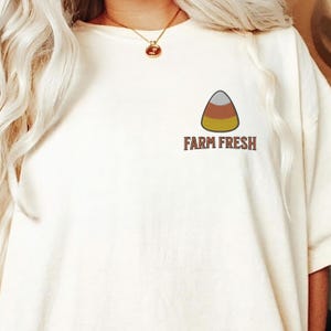 May include: Cream-colored t-shirt featuring a candy corn graphic and the text "FARM FRESH" in brown. The candy corn is a triangle with white, orange, and yellow layers. The shirt has short sleeves and a relaxed fit.