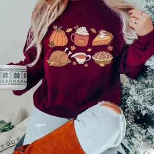 May include: A maroon sweatshirt with a Thanksgiving-themed design featuring a pumpkin, a mug of hot chocolate, a slice of pie, a turkey, gravy, and a pie.