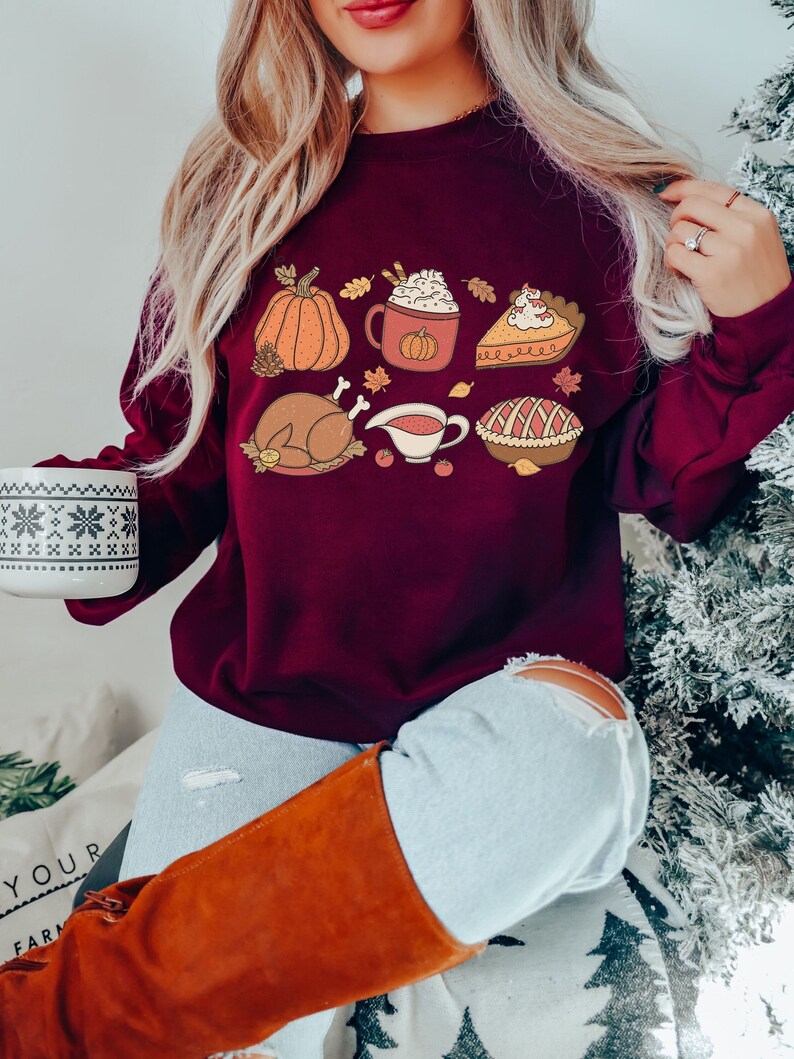 May include: A maroon sweatshirt with a Thanksgiving-themed design featuring a pumpkin, a mug of hot chocolate, a slice of pie, a turkey, gravy, and a pie.