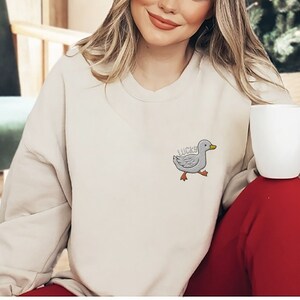 May include: A cream-colored sweatshirt with a gray embroidered duck with the word "LUCKY" on it. The model is wearing red pants and holding a white mug.