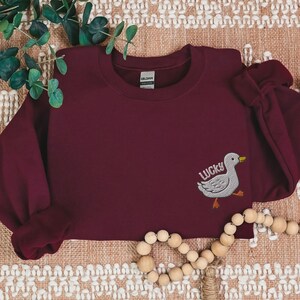 May include: Burgundy sweatshirt with a white embroidered duck and the word "LUCKY" on the left side of the chest.