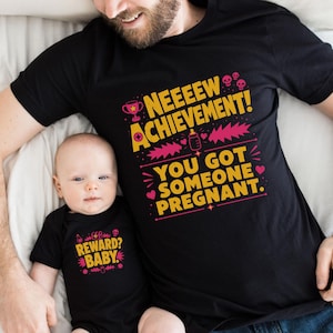 May include: A black short-sleeved t-shirt with yellow and pink text reading NEEEEW ACHIEVEMENT! YOU GOT SOMEONE PREGNANT. and a black baby bodysuit with yellow and pink text reading REWARD? BABY. Both garments feature small graphic icons of skulls, hearts, and a baby bottle.