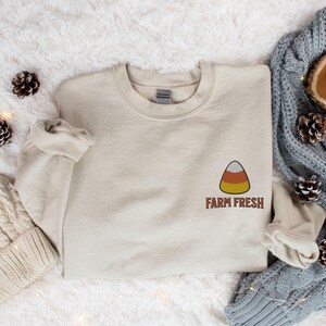 May include: A beige sweatshirt with a small orange, yellow, and brown candy corn graphic and the text "FARM FRESH" in brown.