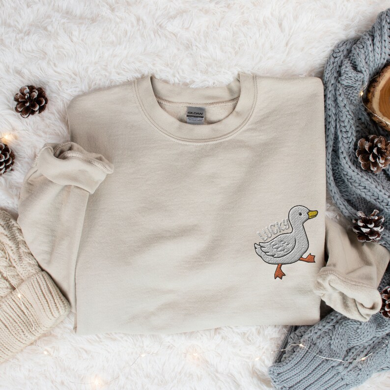 May include: A beige sweatshirt with a white embroidered duck on the left side of the chest. The duck is walking and has the word "lucky" written on it.