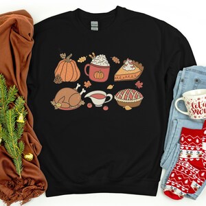 May include: Black sweatshirt with a Thanksgiving-themed design featuring a pumpkin, turkey, pie, gravy, and a cup of hot chocolate.