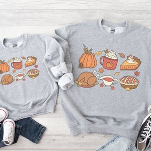 May include: Two gray sweatshirts with a Thanksgiving theme. The design features a turkey, pumpkin, pie, and a cup of coffee with whipped cream. The sweatshirts are laid out on a wooden surface with jeans and sneakers.