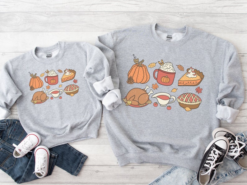 May include: Two gray sweatshirts with a Thanksgiving theme. The design features a turkey, pumpkin, pie, and a cup of coffee with whipped cream. The sweatshirts are laid out on a wooden surface with jeans and sneakers.