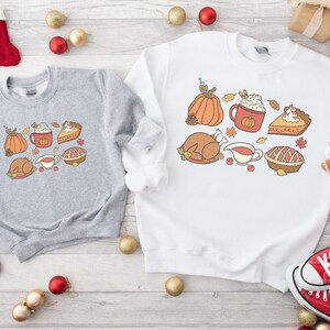 May include: Two gray and white sweatshirts with Thanksgiving-themed graphics. The gray sweatshirt features a turkey, pumpkin, pie, and a cup of coffee with whipped cream. The white sweatshirt features a turkey, pumpkin, pie, and a cup of coffee with whipped cream.