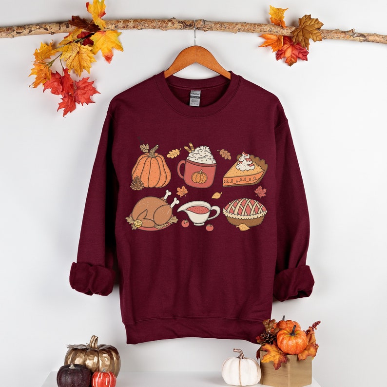 May include: Burgundy sweatshirt with a graphic print of various Thanksgiving foods, including a pumpkin, a turkey, a pie, and a cup of coffee. The graphic is in a vintage style with warm colors.