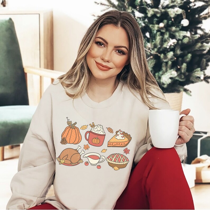 May include: A beige sweatshirt with a Thanksgiving-themed design featuring a pumpkin, a cup of coffee, a turkey, a pie, and a bowl of gravy. The design is in a cartoon style with orange, red, and brown colors.