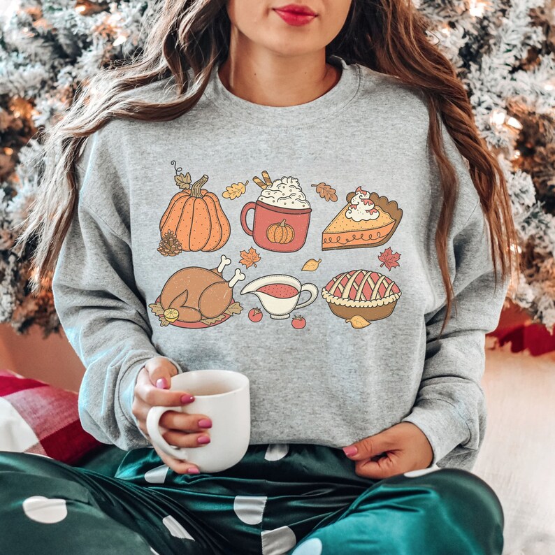 May include: A gray sweatshirt with a Thanksgiving-themed design featuring a pumpkin, a turkey, a pie, a cup of coffee, and a bowl of gravy. The design is in a cartoon style with bright colors.
