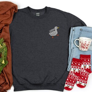 May include: A dark gray sweatshirt with a white embroidered duck on the front. The duck has the word "Lucky" written on it. The sweatshirt is laid out on a white background with a brown scarf, a pair of blue jeans, a white mug with the words "Let it snow" on it, and two red and white Christmas socks.