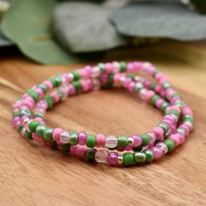 May include: Two beaded bracelets. The bracelets are made of small, round beads in shades of pink, green, and clear. The bracelets are stacked on top of each other, creating a layered effect. The beads are strung on a stretchy cord.