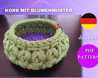 May include: A handmade, green crochet basket with a floral pattern and a purple interior. The basket has a brown leather accent and the text "KORB MIT BLUMENMUSTER" is displayed at the top. The image also includes the German flag and the text "PDF PATTERN".