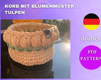May include: A beige and green crocheted basket with a tulip pattern. The basket has a round shape and a leather tag with a button. Text on the image reads "KORB MIT BLUMENMUSTER : TULPEN" and "PDF PATTERN".