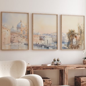 May include: Three framed watercolour paintings depicting cityscapes with buildings, a bridge, and a palm tree. The paintings are hung on a white wall above a wooden console table with a white fluffy armchair in front of it.