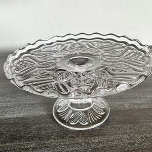 May include: Clear glass cake stand with a scalloped edge and a decorative floral pattern. The stand has a round base with a heart design and a central stem. The cake stand is approximately 10 inches in diameter.