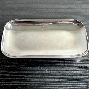 May include: A rectangular, silver-colored tray with rounded corners. The tray has a reflective surface, and the edges are slightly raised. The tray is empty and sits on a dark, textured surface.