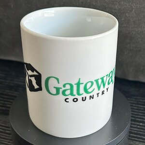 May include: White ceramic mug with the words "Gateway Country" in green and black. The mug has a cylindrical shape and a smooth, glossy finish. The mug is sitting on a dark gray surface.