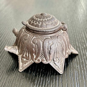 May include: A vintage cast iron inkwell with a hinged lid. The inkwell is decorated with intricate floral patterns and has a star-shaped base.