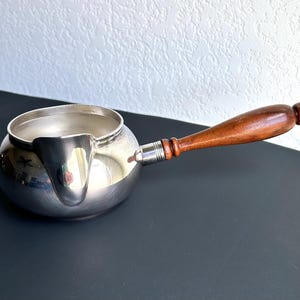 May include: A polished silver-toned metal sauce pot with a rounded body, a wide pouring spout, and a long, tapered wooden handle attached with a silver-colored metal ferrule. The pot features a smooth, reflective surface and a circular opening.