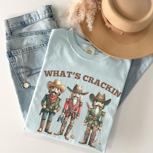 May include: A light blue t-shirt with the phrase "WHAT'S CRACKIN" and a graphic of three cowboy nutcrackers. The shirt is paired with light wash jeans, a tan hat, and a gold necklace.